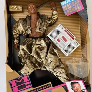 May include: An MC Hammer action figure in a gold metallic jacket and matching pants, packaged in a cardboard box. The set includes a silver boom box, gold-rimmed glasses, and a photo of the artist. Text on the box reads BOOM BOX MAKES REAL RAP SOUNDS.