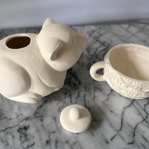 Op de afbeelding: Een crèmekleurige keramische theepot in de vorm van een kat, met een bijpassende kop en deksel. De theepot heeft een grote opening en een gebogen handvat. De kop heeft een getextureerde buitenkant en een handvat. Het deksel is klein en rond met een knop.