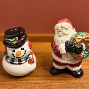 May include: A ceramic snowman and Santa Claus figurine set. The snowman wears a black top hat and a green and red scarf. Santa Claus holds a green wreath with a gold bow. Festive Christmas decor.