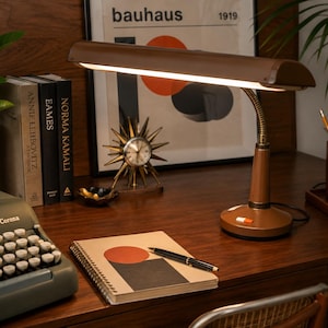 May include: A brown metal desk lamp with a long, rectangular shade and a flexible neck. The lamp features a circular base with a small orange and white power switch. It emits a bright, horizontal beam of light across the workspace surface.