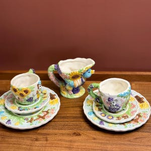 May include: A whimsical ceramic tea set featuring two teacups with matching saucers and a creamer. The cups and saucers are decorated with colorful floral and animal designs. The creamer has a unique handle and a similar floral pattern.