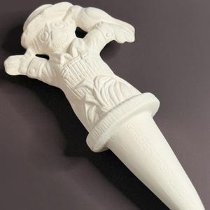 May include: A white ceramic bottle stopper in the shape of a scarecrow. The scarecrow has a hat, arms outstretched, and a smiling face. The stopper has a conical shape with the text "@ALBERTASSTUDIO" printed on it.