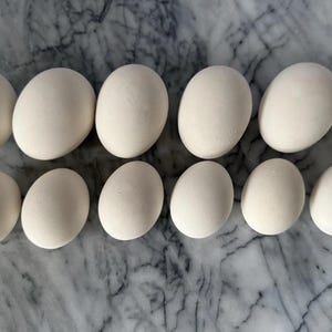 May include: A dozen white eggs arranged in two rows on a marble surface. The eggs have a smooth, oval shape and a creamy white color. The background is a gray and white marble pattern.