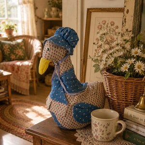 May include: A fabric goose figurine featuring a beige body with a small red floral pattern, a bright yellow beak, and a blue bonnet with a white floral print. The goose wears a matching blue capelet tied with a light purple ribbon around its neck.
