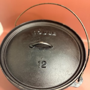 Peut inclure: Une cocotte en fonte noire avec un couvercle et une poignée en fil de fer. Le couvercle porte le numéro 12 en relief. Le pot est conçu pour la cuisine et est probablement utilisé pour le camping ou la cuisine en plein air.