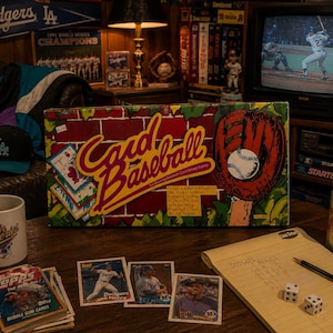 Könnte beinhalten: Eine Card Baseball Brettspielschachtel mit rotem Backsteinmuster, einem Baseballhandschuh und einem Ball. Die Schachtel trägt die Aufschrift Card Baseball und The Real Baseball Card Board Game. Drumherum liegen Baseballkarten, ein Becher, ein Notizblock, Würfel und eine Diet Coke Dose.