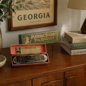 May include: A silver M. Hohner Marine Band harmonica rests inside a pink and yellow cardboard box. The box lid features a vintage paper label with text including Marine Band Full Concert and M. Hohner. The harmonica has engraved metal plates.
