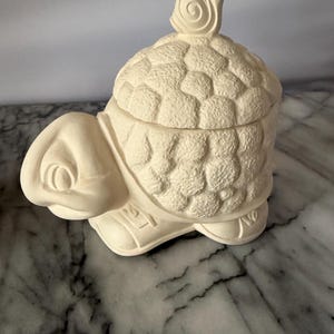 Może przedstawiać: Ceramiczny pojemnik w kształcie żółwia z teksturowaną skorupą i uchwytem w kształcie ślimaka na pokrywie. Pojemnik jest w kolorze złamanej bieli i stoi na marmurowej powierzchni.