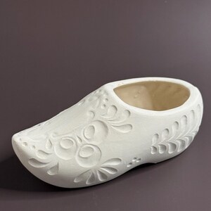 May include: A white ceramic clog-shaped planter with floral relief detailing. The planter has an open top and is designed to hold small plants or flowers. The decorative elements include stylized flowers and leaves, adding a touch of rustic charm.