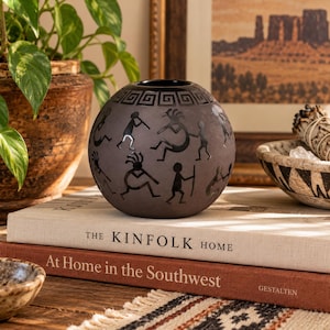 May include: A spherical dark gray ceramic vessel featuring a black Greek key pattern around the top rim and black silhouettes of flute-playing figures around the body. The object is resting on a stack of two books titled The Kinfolk Home.