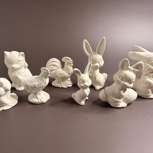 Può includere: Una collezione di statuette in ceramica non dipinte, tra cui un gatto, un gallo, una rana e diversi conigli. Le statuette sono in varie pose e dimensioni, perfette per progetti di pittura fai-da-te. Lo sfondo è di colore neutro.