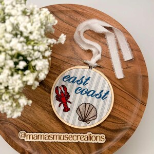 May include: An embroidered hoop art piece with the words "east coast" in blue script. The design includes a red lobster and a brown seashell on a striped background. A white ribbon is attached for hanging. The piece is displayed on a wooden tray.
