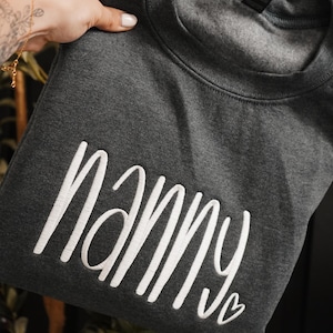 May include: A dark charcoal gray crewneck sweatshirt featuring the word nanny embroidered in white cursive script across the front, accompanied by a small white embroidered heart outline at the end of the text.