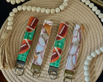 May include: Four fabric key fobs with metal hardware. Two fobs feature a football design on a green field, and two feature baseballs. The fobs are on a woven beige placemat with decorative wooden beads.