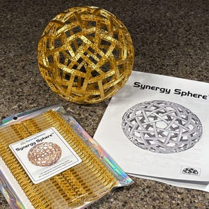 May include: A gold-colored Synergy Sphere puzzle constructed from interlocking pieces, accompanied by a clear plastic bag containing additional gold components and a white instruction manual featuring the Synergy Sphere text and illustration.