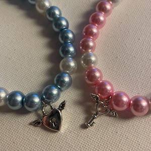 May include: Two beaded bracelets, one blue and one pink, with white accent beads. Each bracelet has a silver charm, one a heart with wings and the other a key with wings. The bracelets are on a white surface.