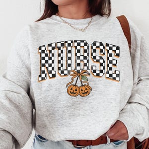 Puede incluir: Una sudadera de cuello redondo en gris jaspeado claro con la palabra NURSE en un patr&oacute;n de cuadros blanco y negro. Debajo del texto hay dos calabazas naranjas unidas por un lazo, que recuerdan a un par de cerezas.