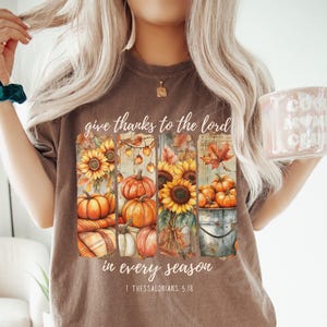 May include: Espresso-colored t-shirt with a fall-themed graphic. The graphic features sunflowers, pumpkins, and the text "give thanks to the lord in every season" and "1 Thessalonians 5:18". The shirt is a comfortable, casual style.