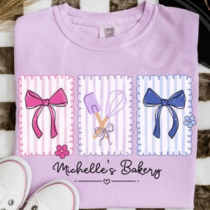 May include: Orchid-colored t-shirt with a bakery-themed design. The design includes three framed illustrations of a pink bow, a spatula and whisk, and a blue bow. The text "Michelle's Bakery" is below the illustrations. White sneakers are at the bottom.
