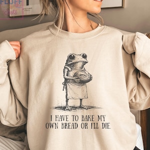 May include: A sand-colored crewneck sweatshirt featuring a black ink-style illustration of a frog wearing an apron and holding a loaf of bread. Below the graphic, the text reads: I HAVE TO BAKE MY OWN BREAD OR I'LL DIE. The garment has long sleeves.