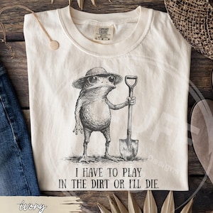 May include: Ivory-colored t-shirt featuring a detailed illustration of a frog wearing a hat and holding a shovel. The text "I HAVE TO PLAY IN THE DIRT OR I'LL DIE" is printed below the frog. The shirt is a Comfort Colors brand.