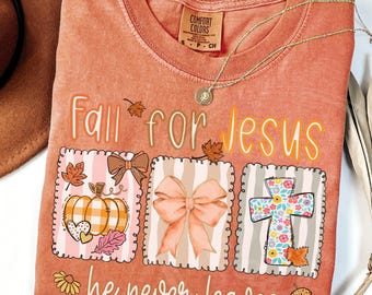 May include: A terracotta-colored t-shirt with a crew neckline and a graphic print. The text reads Fall for Jesus he never leaves. The design features three rectangular panels containing a pumpkin, a bow, and a floral cross with autumn leaves.