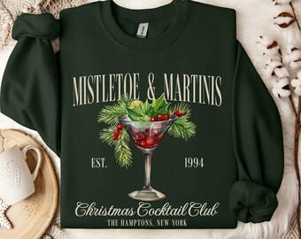 May include: A dark green long-sleeve sweatshirt featuring a graphic of a martini glass filled with red berries and pine needles. The text reads MISTLETOE & MARTINIS, EST. 1994, Christmas Cocktail Club, and THE HAMPTONS, NEW YORK in a cream-colored font.