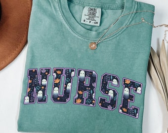 May include: A seafoam green t-shirt with the word "NURSE" in a collegiate style font. The letters are filled with a floral and ghost pattern. A gold necklace with a pendant is draped across the shirt.