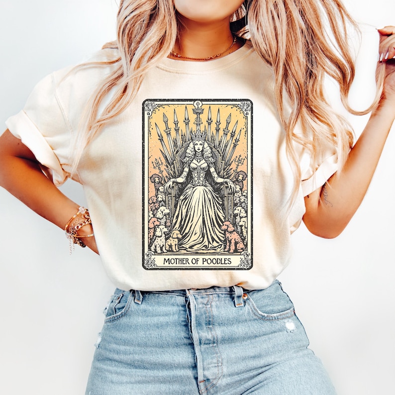 May include: Cream-colored t-shirt featuring a tarot card design with a queen figure on a throne surrounded by poodles. The text "MOTHER OF POODLES" is at the bottom of the card. The shirt is paired with blue jeans.