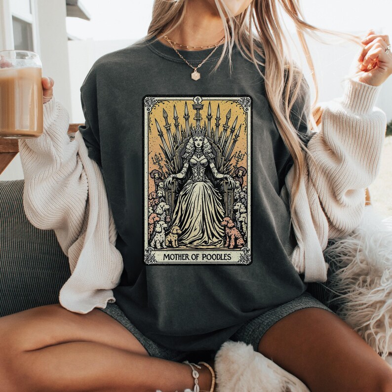 May include: Dark gray sweatshirt featuring a tarot card design with a queen seated on a throne, surrounded by poodles. The text "MOTHER OF POODLES" is at the bottom of the card. The design is in shades of gold, cream, and black.