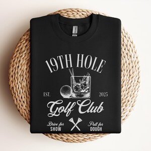 K&ouml;nnte beinhalten: Schwarzes T-Shirt mit wei&szlig;em Text, der "19th Hole Golf Club Est. 2025" lautet, mit einem Golfball, Golfschl&auml;gern und einem Glas Eis mit einem Strohhalm.