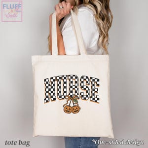Puede incluir: Un bolso de tela color crema con la palabra NURSE en un patr&oacute;n de cuadros blanco y negro. Debajo del texto, hay un gr&aacute;fico de dos calabazas naranjas tipo jack-o'-lantern conectadas por un tallo con una hoja verde y un lazo marr&oacute;n.