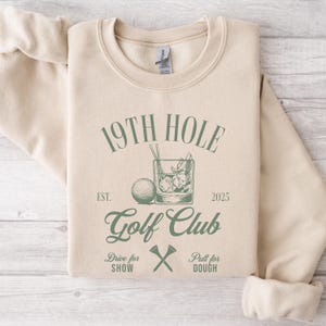 K&ouml;nnte beinhalten: Ein beiger Pullover mit einem gr&uuml;nen Grafikdesign, auf dem "19th Hole Golf Club Est. 2025" mit einem Golfball und einem Glas Eis steht. Der Text unter der Grafik lautet "Drive for Show" und "Putt for Dough" mit einem gekreuzten Satz Golfclubs.