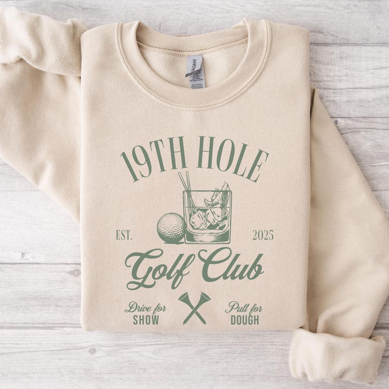 K&ouml;nnte beinhalten: Ein beiger Pullover mit einem gr&uuml;nen Grafikdesign, auf dem "19th Hole Golf Club Est. 2025" mit einem Golfball und einem Glas Eis steht. Der Text unter der Grafik lautet "Drive for Show" und "Putt for Dough" mit einem gekreuzten Satz Golfclubs.