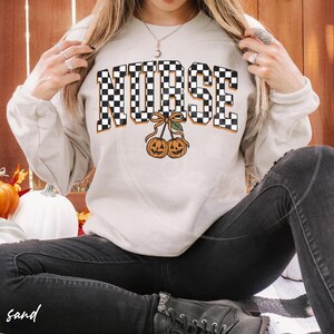 Puede incluir: Una sudadera de cuello redondo color arena con la palabra NURSE en un patr&oacute;n de cuadros blanco y negro. Debajo del texto hay dos calabazas naranjas que parecen cerezas, unidas por un tallo marr&oacute;n y una peque&ntilde;a hoja verde.