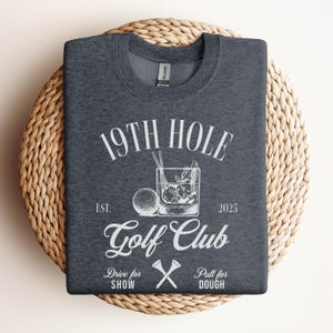 K&ouml;nnte beinhalten: Ein dunkelgraues T-Shirt mit einem wei&szlig;en Grafikdesign, auf dem "19th Hole Golf Club Est. 2025" mit einem Golfball, einem Glas Eis und gekreuzten Golfschl&auml;gern steht. Der Text unter dem Logo lautet "Drive for Show Pull for Dough".
