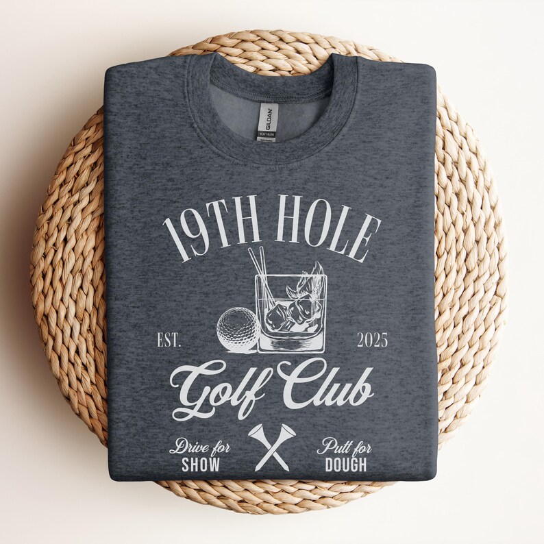 K&ouml;nnte beinhalten: Ein dunkelgraues T-Shirt mit einem wei&szlig;en Grafikdesign, auf dem "19th Hole Golf Club Est. 2025" mit einem Golfball, einem Glas Eis und gekreuzten Golfschl&auml;gern steht. Der Text unter dem Logo lautet "Drive for Show Pull for Dough".