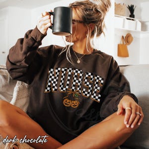 Puede incluir: Una sudadera de manga larga en color marr&oacute;n chocolate oscuro con la palabra NURSE en un patr&oacute;n de cuadros blanco y negro. Debajo del texto, hay un gr&aacute;fico de dos calabazas de Halloween naranjas atadas con un lazo, parecidas a cerezas.