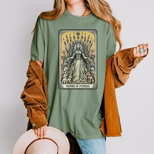 May include: Sage green t-shirt featuring a tarot card design. The card depicts a queen seated on a throne, surrounded by poodles, with the text "MOTHER OF POODLES" at the bottom. The queen wears a crown and a long gown.