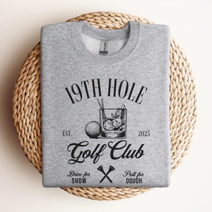 K&ouml;nnte beinhalten: Grauer Pullover mit einem schwarzen grafischen Design, auf dem "19th Hole Golf Club Est. 2025" mit einem Golfball und einem Getr&auml;nk steht. Der Text darunter lautet "Drive for Show Pull for Dough" mit einem Paar gekreuzter Golfschl&auml;ger.