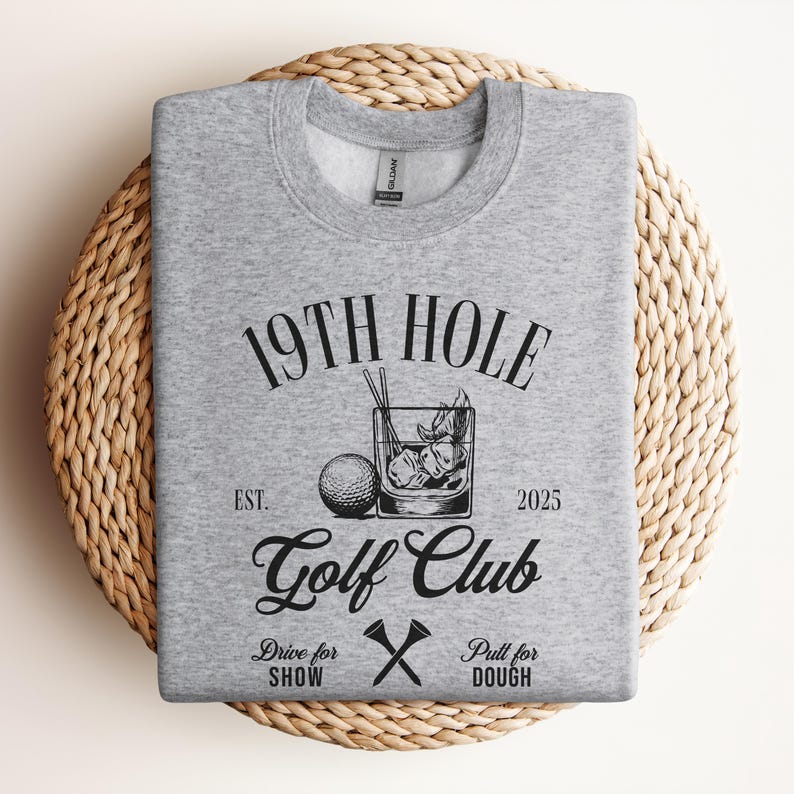 K&ouml;nnte beinhalten: Grauer Pullover mit einem schwarzen grafischen Design, auf dem "19th Hole Golf Club Est. 2025" mit einem Golfball und einem Getr&auml;nk steht. Der Text darunter lautet "Drive for Show Pull for Dough" mit einem Paar gekreuzter Golfschl&auml;ger.