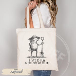 May include: A natural canvas tote bag featuring a whimsical illustration of a frog wearing a hat and holding a shovel. The text "I HAVE TO PLAY IN THE DIRT OR I'LL DIE" is printed below the frog. The word "natural" is printed at the bottom.