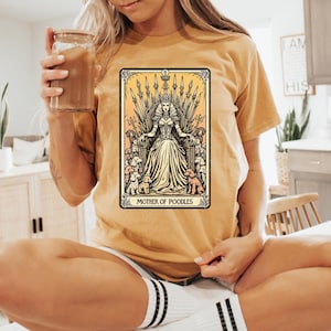 May include: Mustard yellow t-shirt featuring a tarot card-style graphic. The graphic depicts a woman on a throne surrounded by poodles, with the text "MOTHER OF POODLES" at the bottom. The shirt is a relaxed fit.