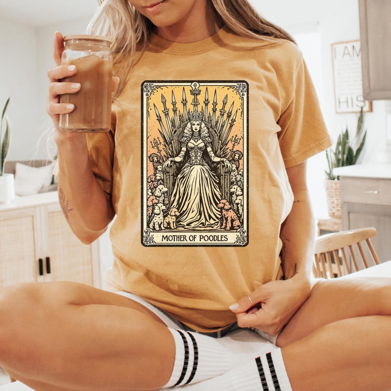 May include: Mustard yellow t-shirt featuring a tarot card-style graphic. The graphic depicts a woman on a throne surrounded by poodles, with the text "MOTHER OF POODLES" at the bottom. The shirt is a relaxed fit.
