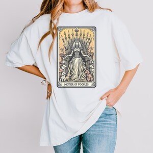 May include: Cream-colored t-shirt featuring a tarot card-style graphic. The graphic depicts a queen seated on a throne, surrounded by poodles, with the text "MOTHER OF POODLES" at the bottom. The design is in black and cream tones.