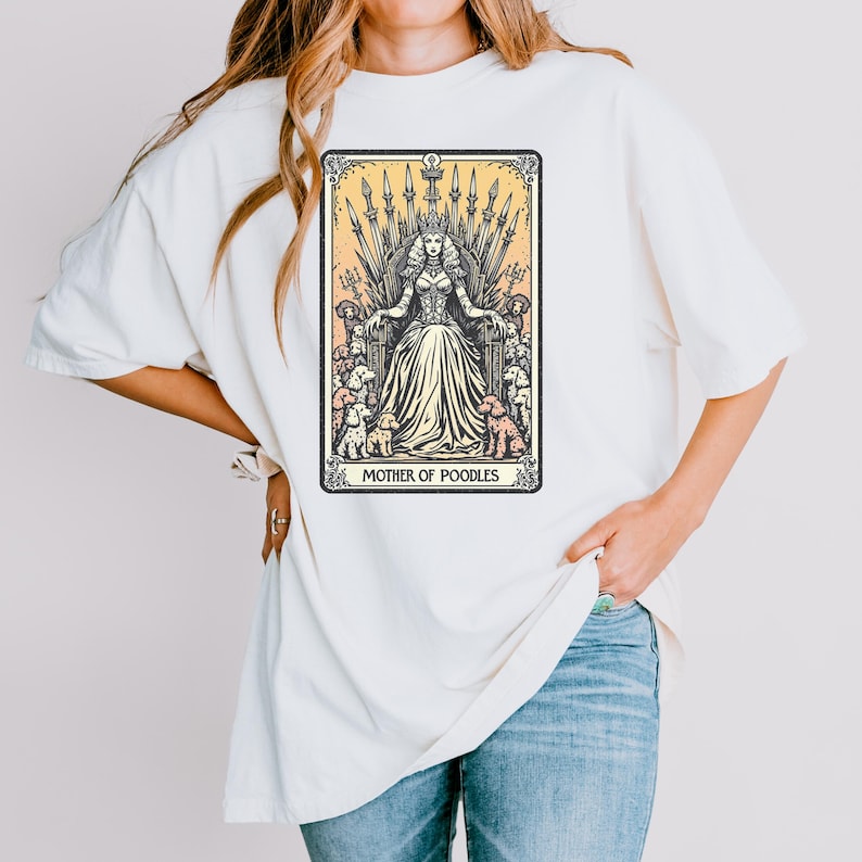 May include: Cream-colored t-shirt featuring a tarot card-style graphic. The graphic depicts a queen seated on a throne, surrounded by poodles, with the text "MOTHER OF POODLES" at the bottom. The design is in black and cream tones.