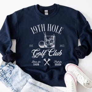 K&ouml;nnte beinhalten: Ein marineblauer Sweatshirt mit wei&szlig;em Text, der "19th Hole Golf Club Est. 2025" lautet, mit einem Golfball und einem Glas Eis mit einem Strohhalm. Der Text unter dem Logo lautet "Drive for Show Putt for Dough".