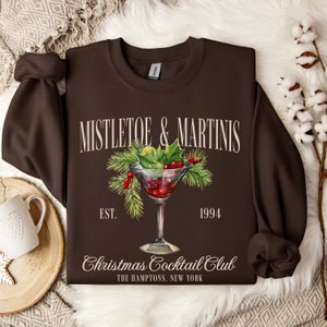 May include: A dark brown crewneck sweatshirt featuring a graphic of a martini glass filled with red berries and garnished with pine needles and holly leaves. The text reads MISTLETOE & MARTINIS, EST. 1994, Christmas Cocktail Club, THE HAMPTONS, NEW YORK.