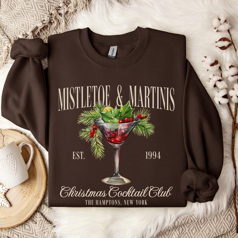May include: A dark brown crewneck sweatshirt featuring a graphic of a martini glass filled with red berries and garnished with pine needles and holly leaves. The text reads MISTLETOE & MARTINIS, EST. 1994, Christmas Cocktail Club, THE HAMPTONS, NEW YORK.