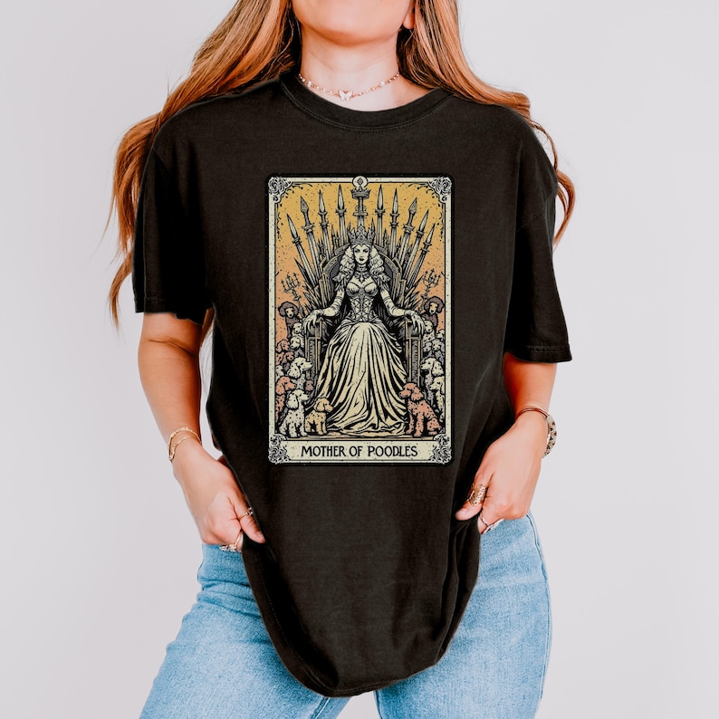 May include: Black t-shirt featuring a tarot card design with a woman on a throne surrounded by poodles. The card reads "MOTHER OF POODLES" in a decorative frame with a yellow and orange background.